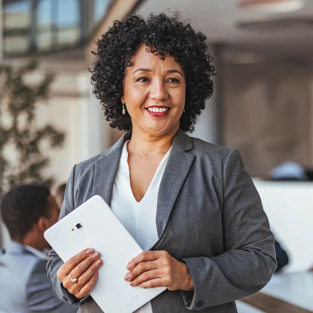 Professional woman in a business suit holding a tablet, smiling confidently in a modern office setting.